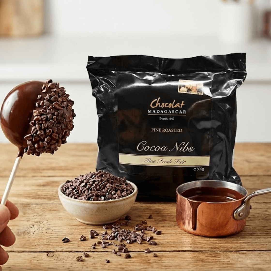 Chocolat Madagascar Fine Roasted Cocoa Nibs in a black 500 g package on a wooden table, with a bowl of cocoa nibs, melted dark chocolate in a small saucepan, and a hand holding a cake pop half coated in chocolate and cocoa nibs in a clean kitchen setting.